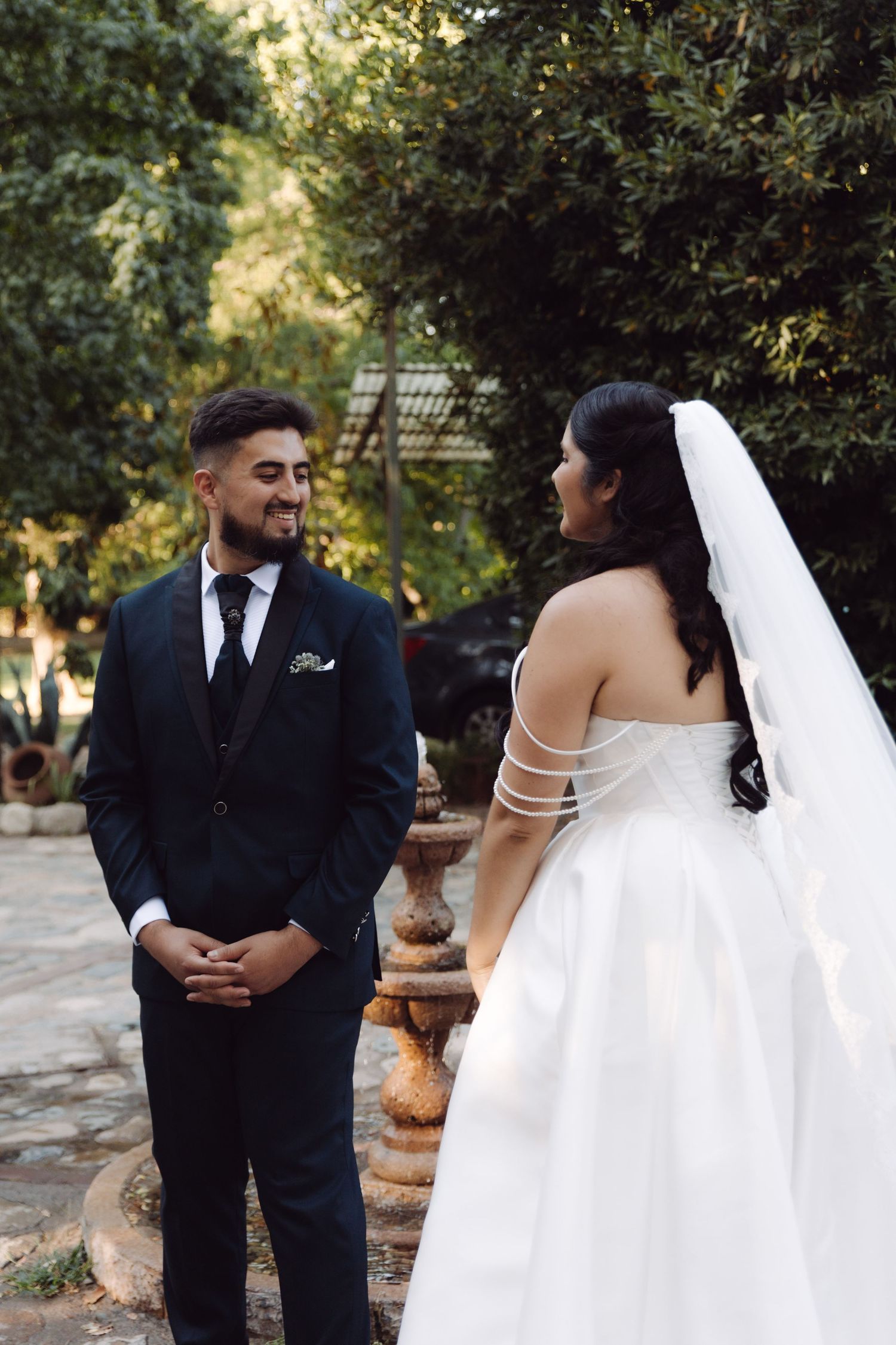 Groom in navy suit smiles warmly at bride in strapless white wedding gown with pearl straps in garden.