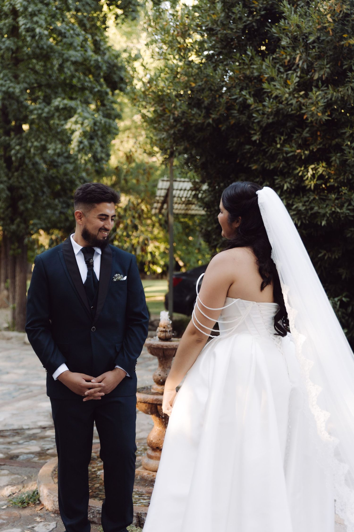 Groom clasps hands smiling at bride in white strapless ball gown with long veil during outdoor wedding photos.