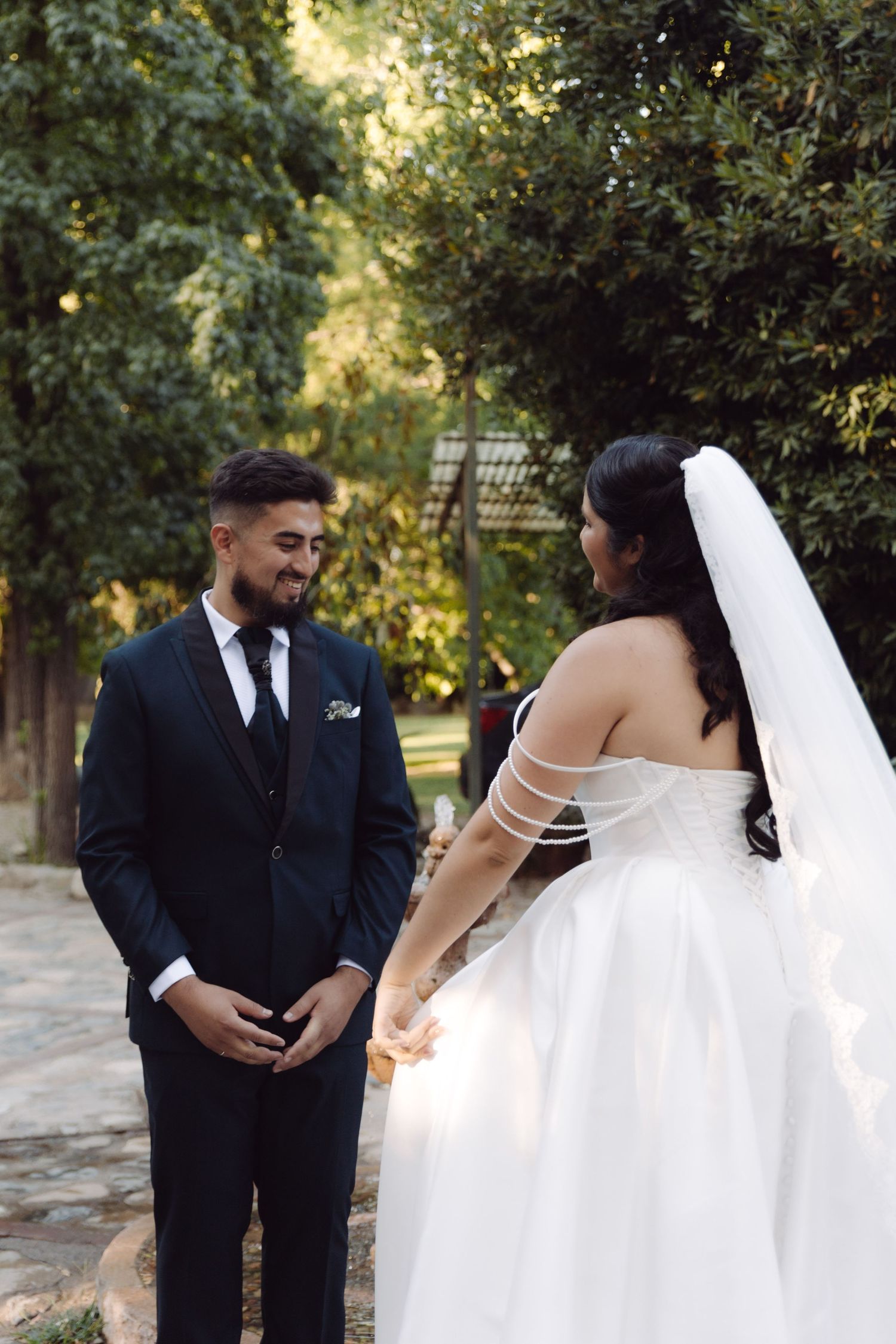 Groom in navy suit smiles holding brides hand as she stands in white strapless gown in sunlit garden.
