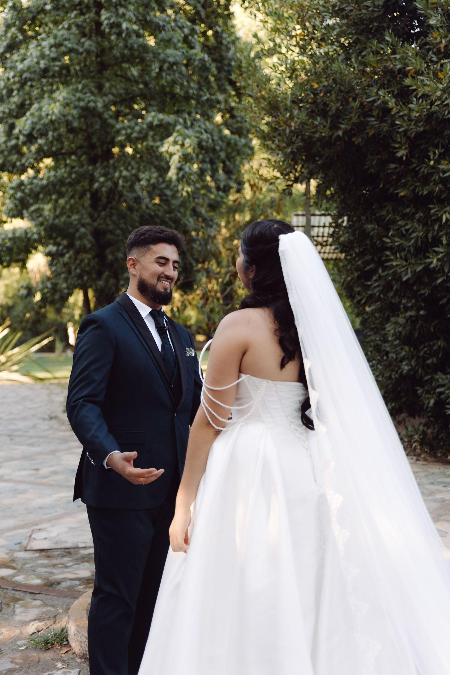 Groom in navy suit smiles tenderly at bride in white strapless ball gown with long veil in garden setting.