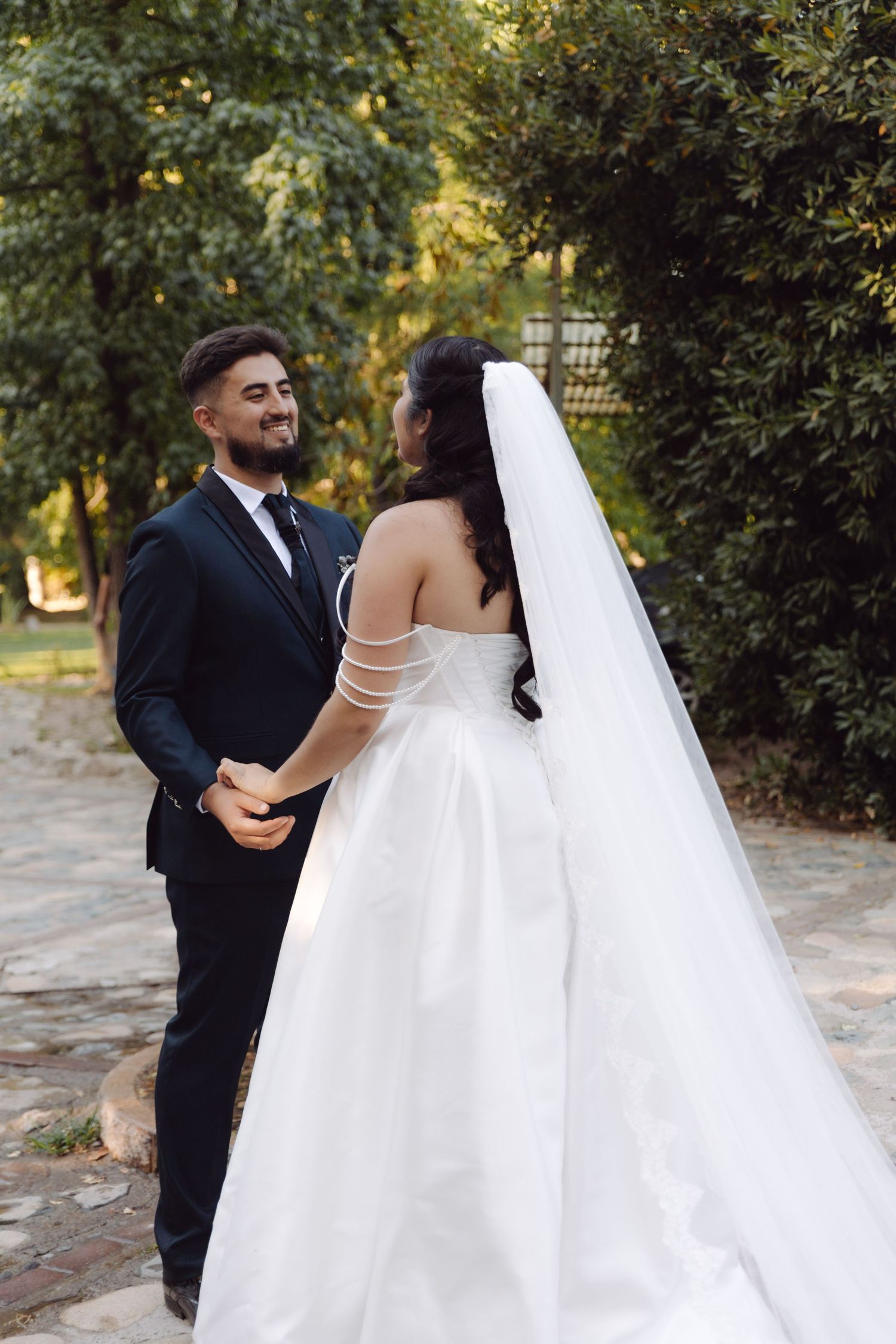 Bride in white off-shoulder gown and veil holds hands with smiling groom in black suit outdoors.