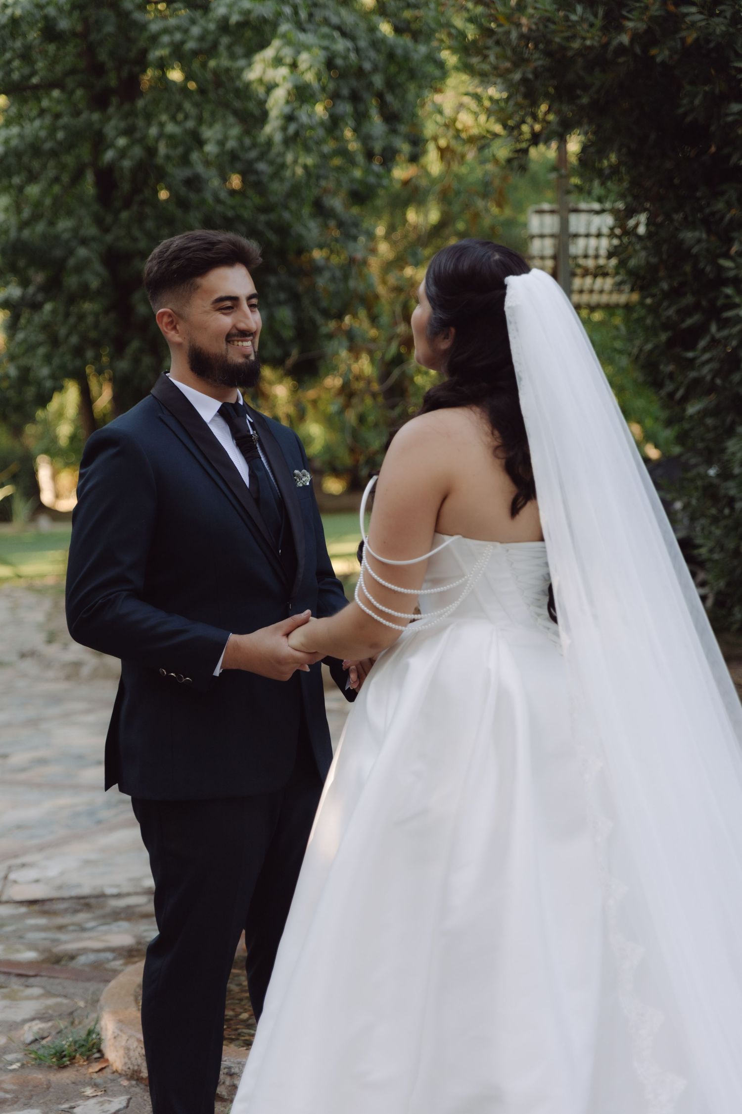 Happy groom in dark suit smiles at bride in white wedding dress and veil in a garden setting.