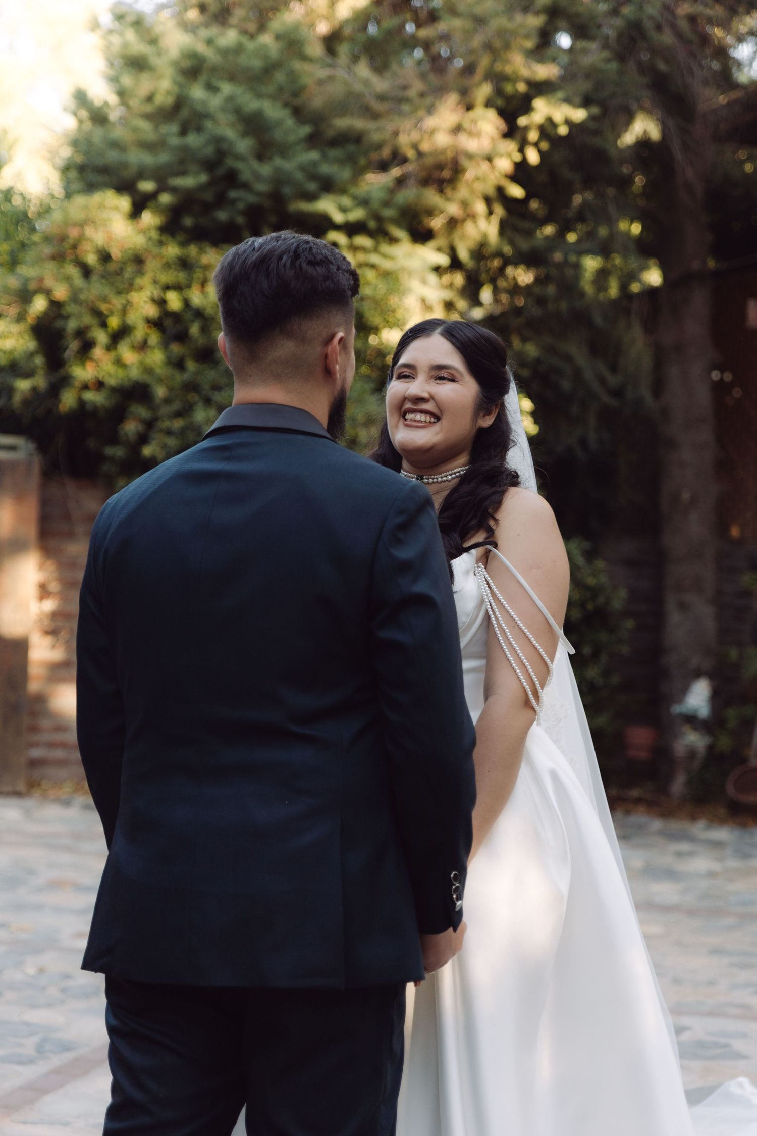 Smiling bride in white satin gown with strappy back detail faces groom in black suit outdoors.