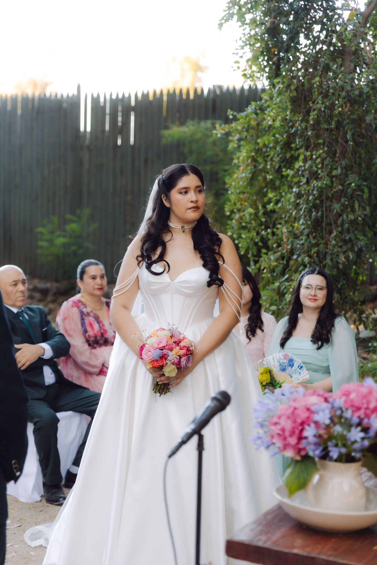Elegant bride in white sweetheart gown holds colorful bouquet at outdoor wedding ceremony altar.