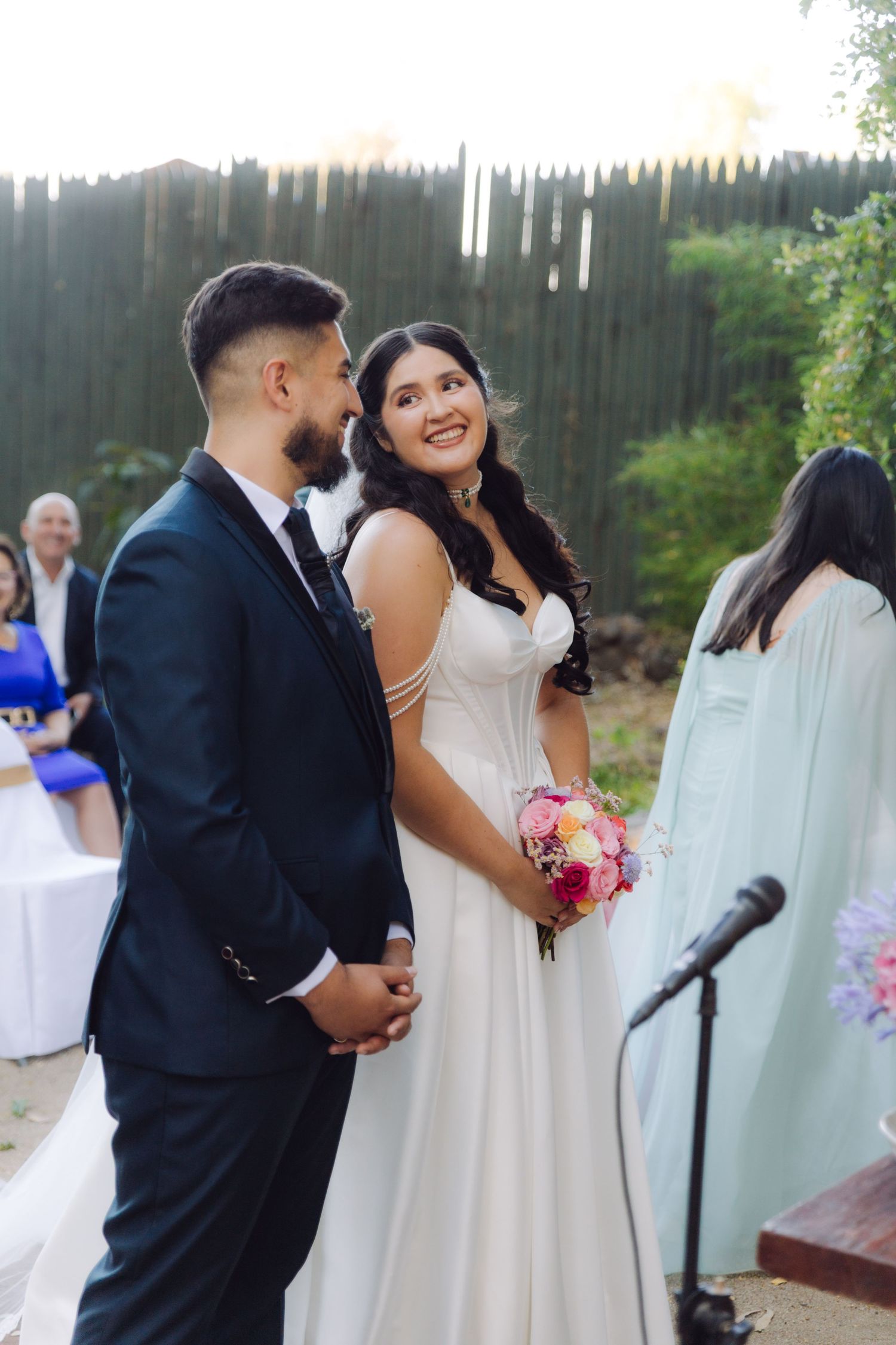 Bride smiling at groom during outdoor wedding ceremony, holding colorful bouquet in white gown.