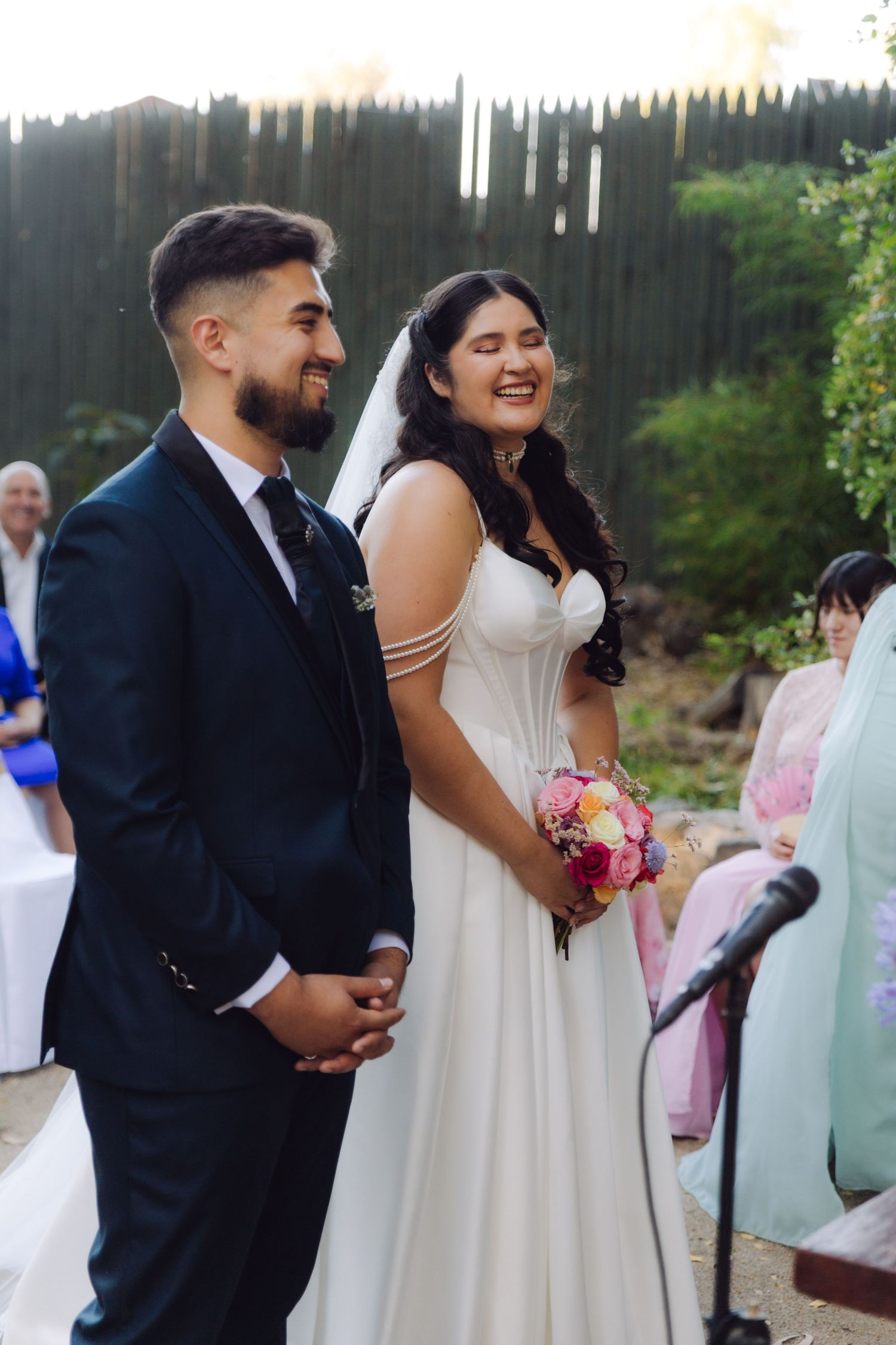 Happy bride and groom laughing together at outdoor wedding ceremony with colorful bouquet.