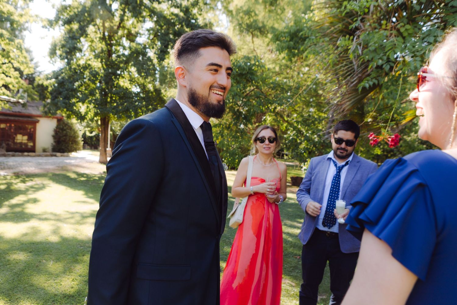 Smiling groom in a dark suit and tie socializing with guests outdoors at a sunny wedding reception.