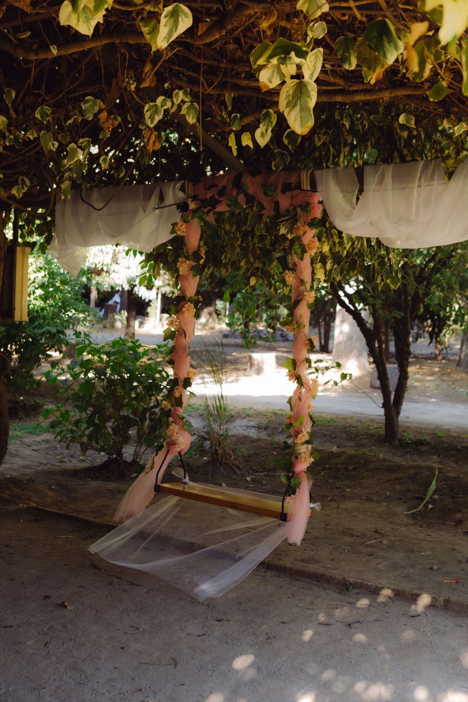 Decorative floral swing adorned with pink flowers and white fabric hanging under a leafy pergola outdoors.