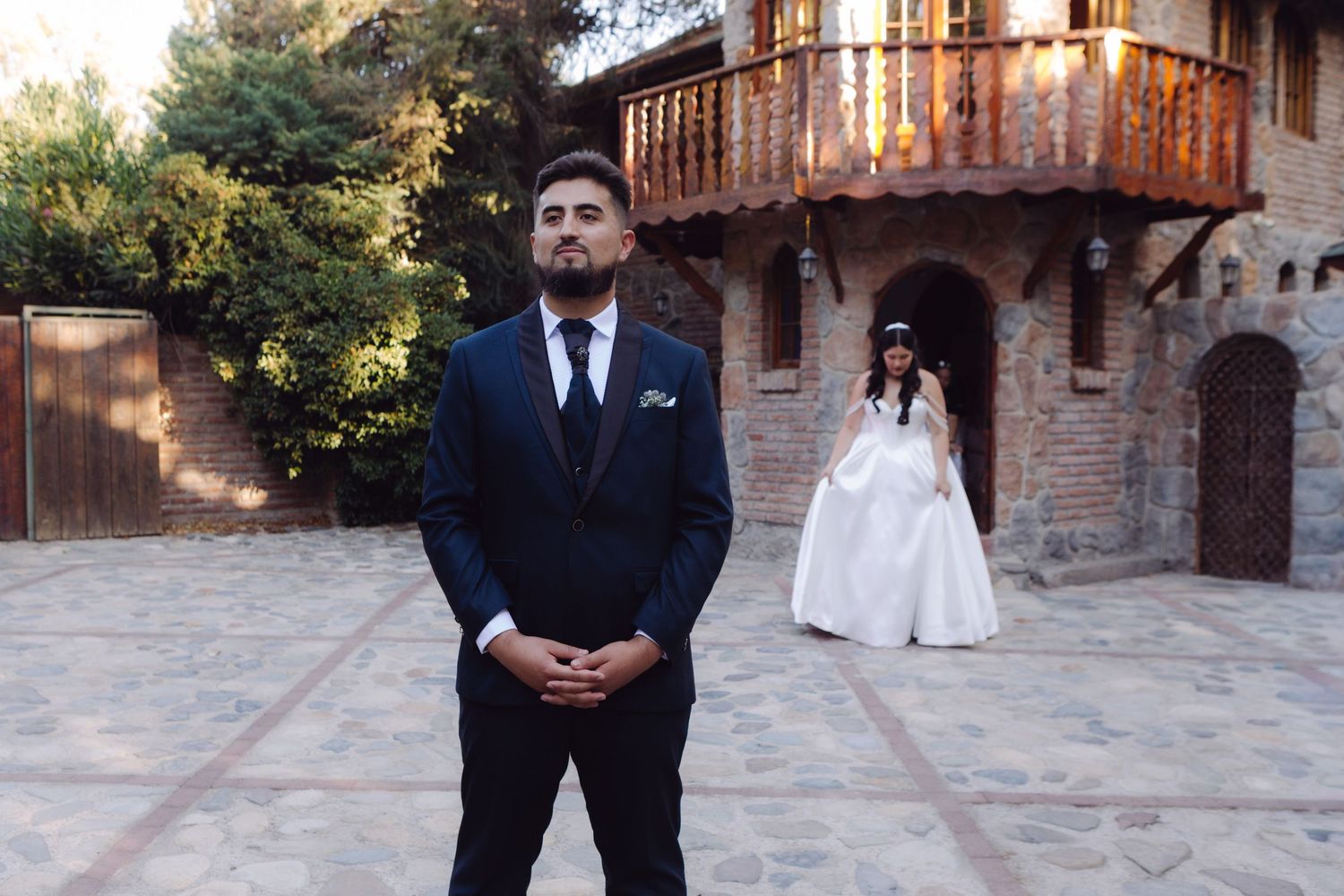 Groom in dark suit standing in foreground as bride in white gown approaches at a rustic brick venue.