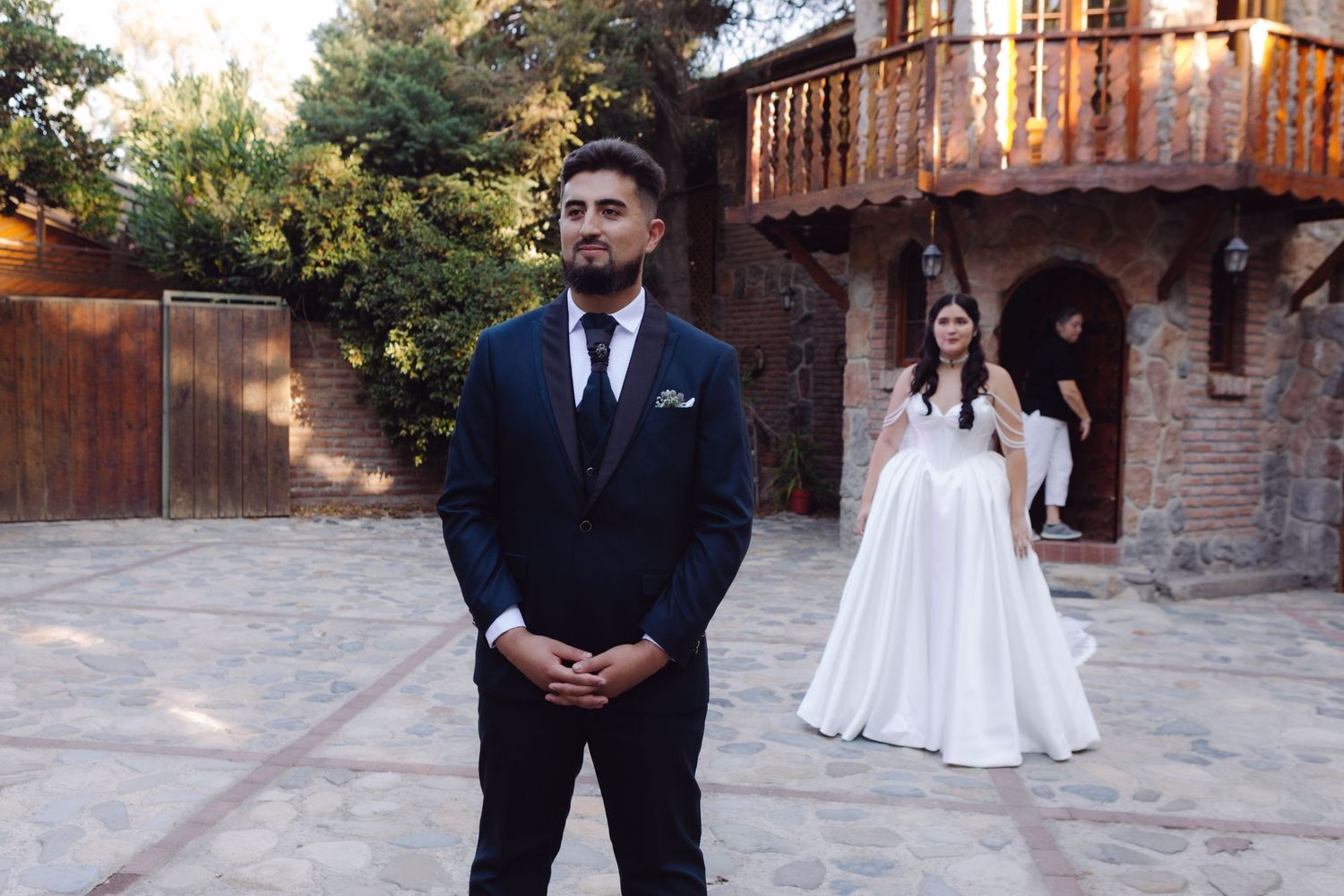 Bride in a flowing white wedding gown walking toward groom in a dark suit at a rustic outdoor venue.