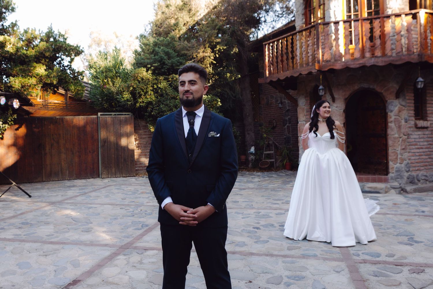 Groom in navy suit stands in courtyard as bride in white ball gown approaches for first look at outdoor wedding venue.