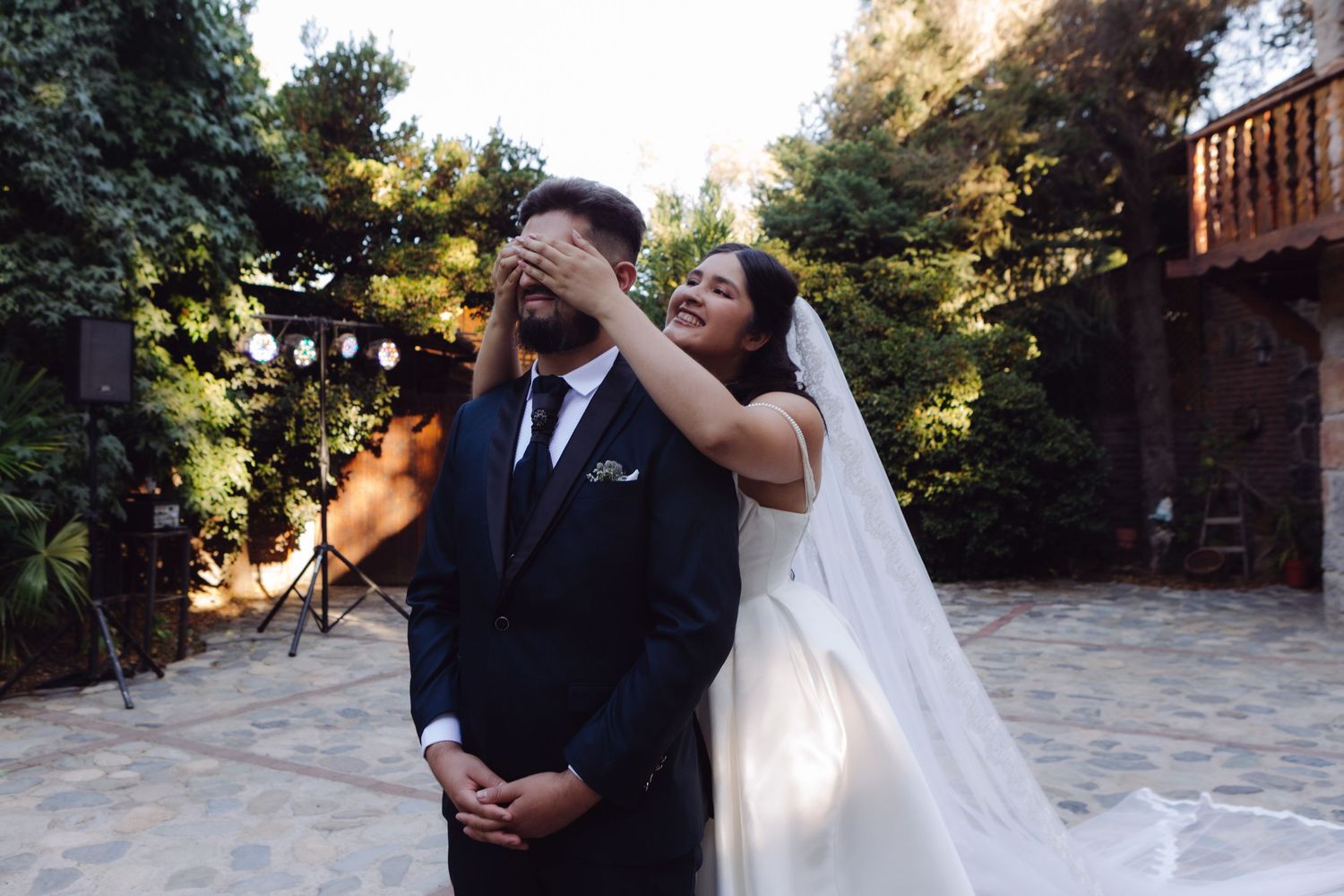 Bride in white wedding dress covers grooms eyes with her hands during romantic first look moment outdoors.