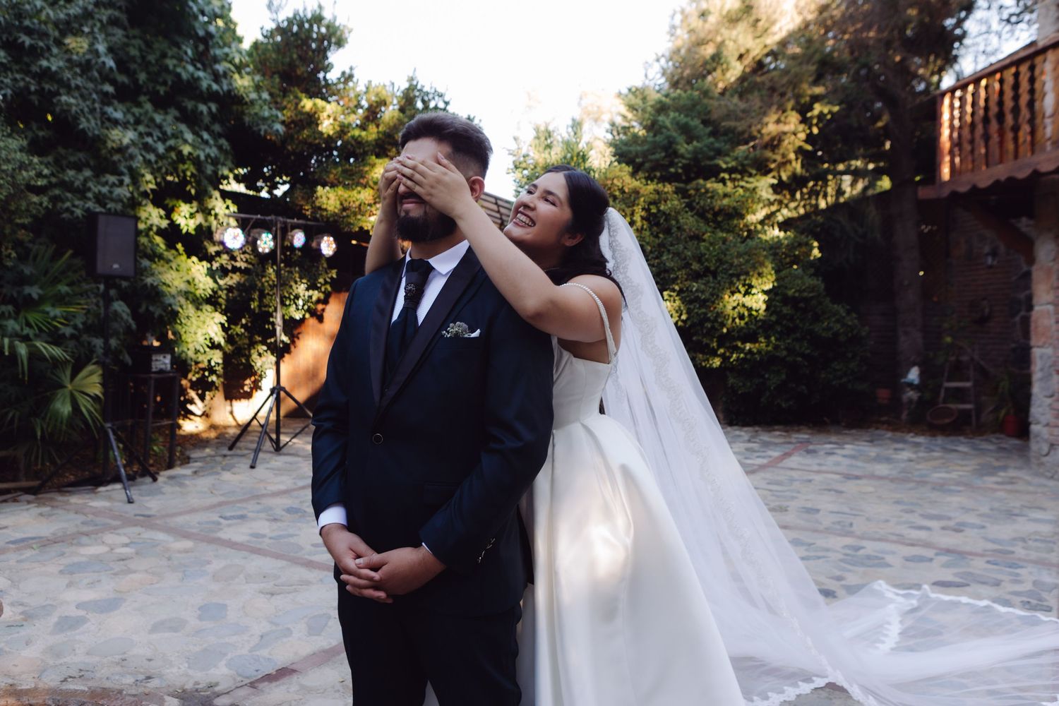 Smiling bride in white veil covers grooms eyes with both hands during wedding first look in garden courtyard.