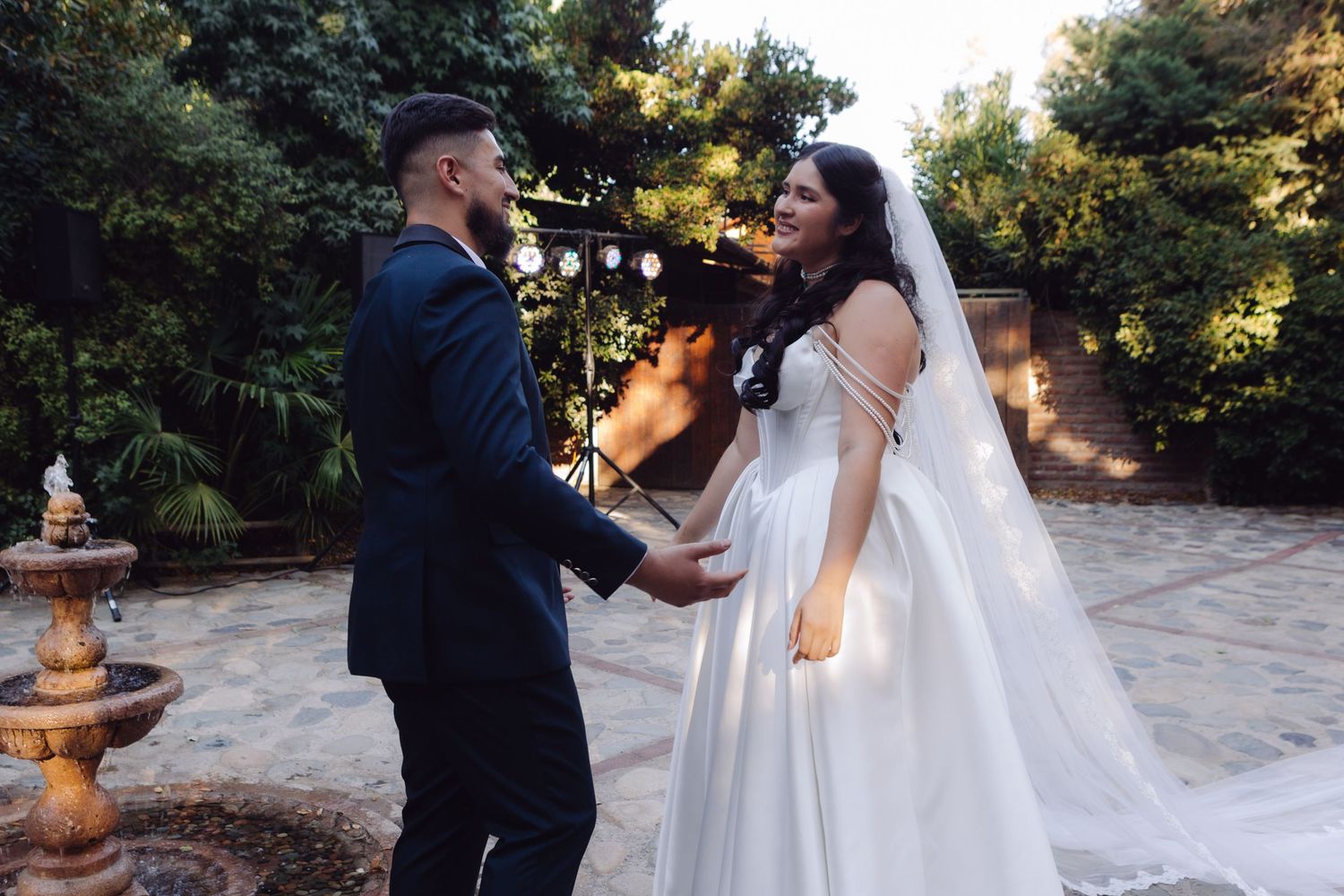 Bride and groom share joyful first look moment holding hands in lush garden with fountain at outdoor wedding.