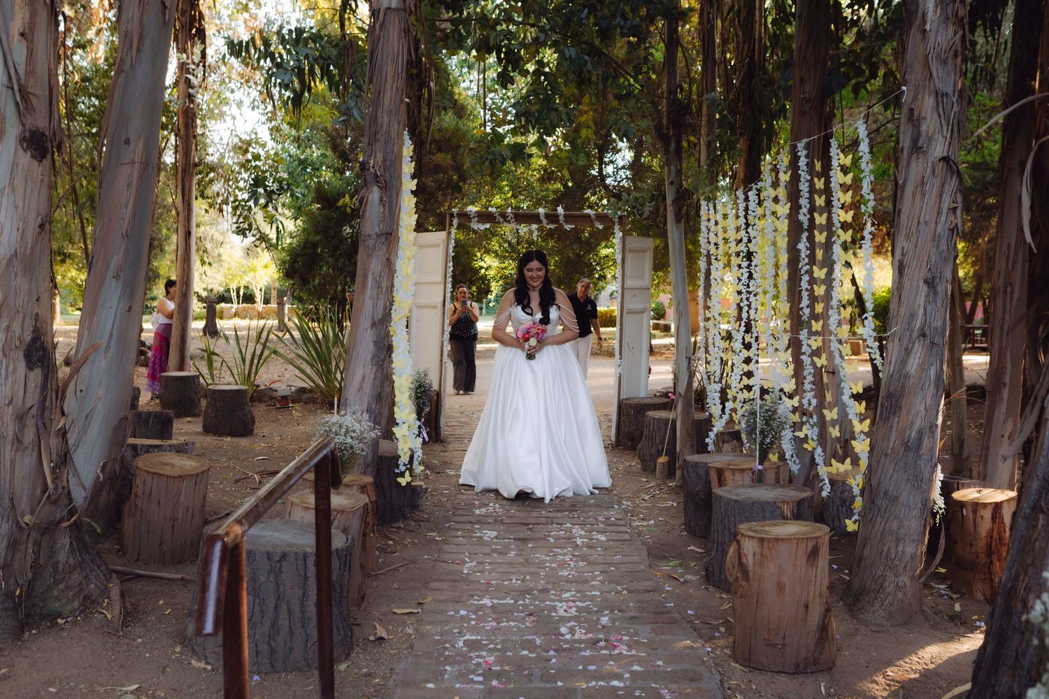 Bride in white gown walks down a tree-lined outdoor aisle decorated with floral garlands.