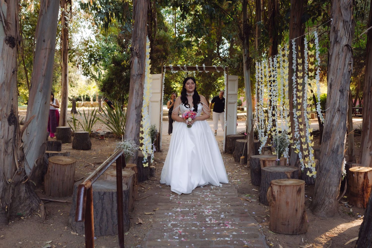Bride holding bouquet walks through a rustic outdoor wedding aisle flanked by tall trees.