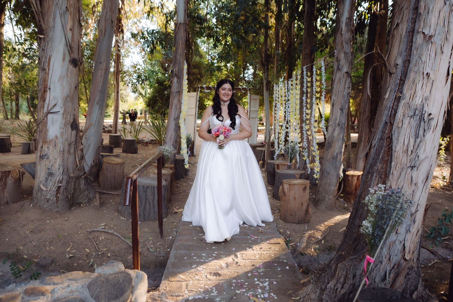 Bride in flowing white dress walks through a sunlit outdoor venue surrounded by trees and cacti.
