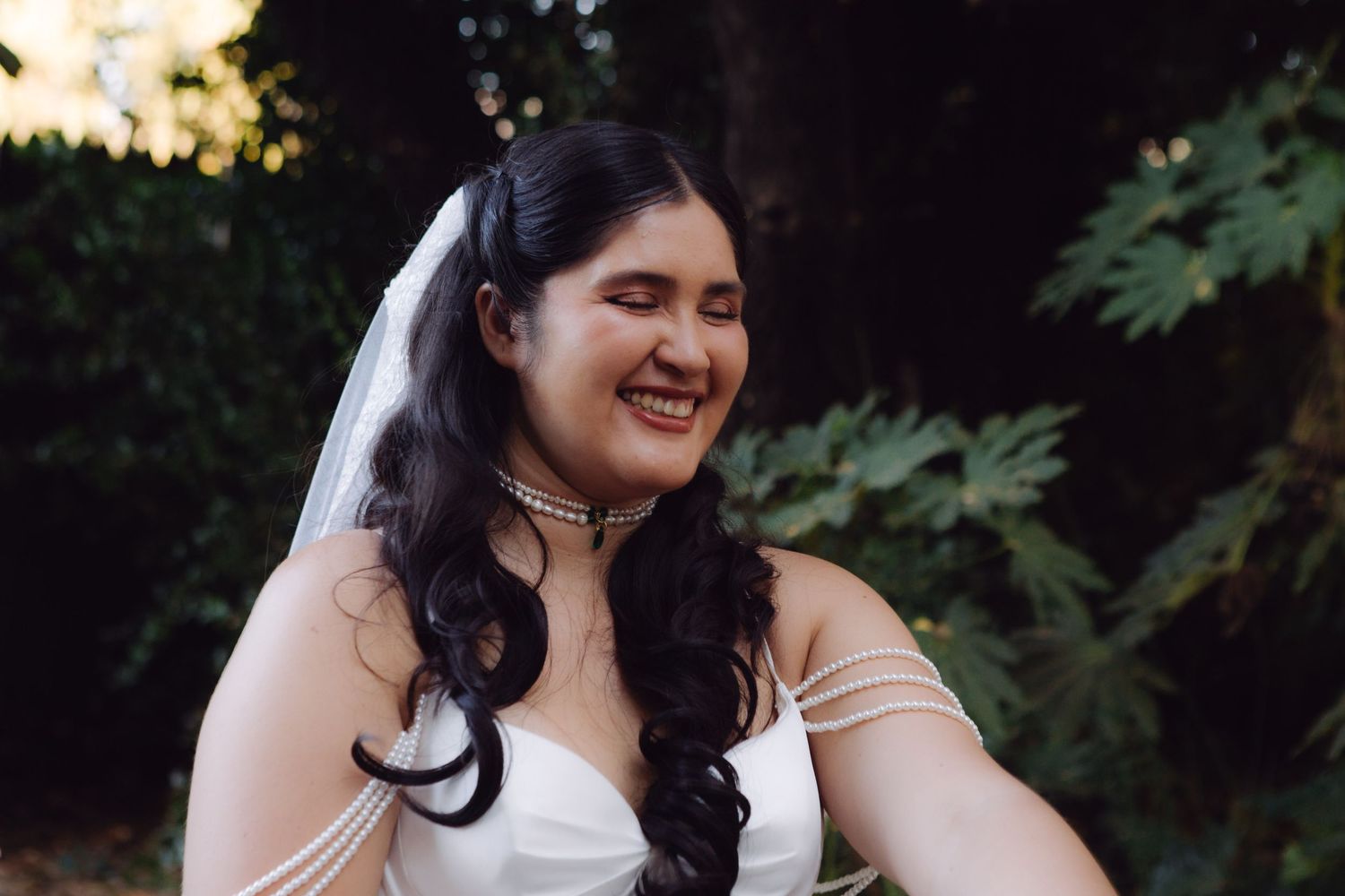 Smiling bride in white off-shoulder gown and veil poses outdoors with lush green foliage behind her.