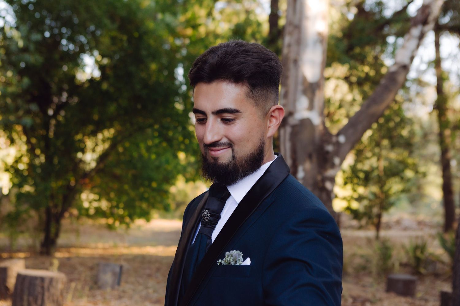 Handsome groom in navy suit with tie and pocket square smiles softly in an outdoor setting.