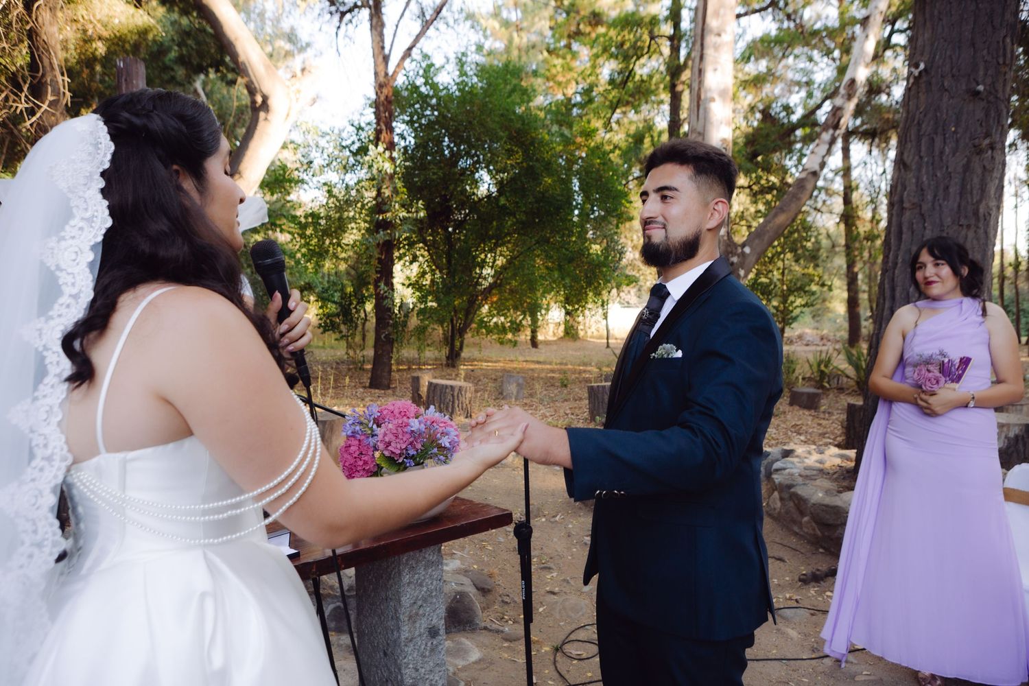 Bride and groom exchange vows at an outdoor woodland wedding ceremony with bridesmaid nearby.