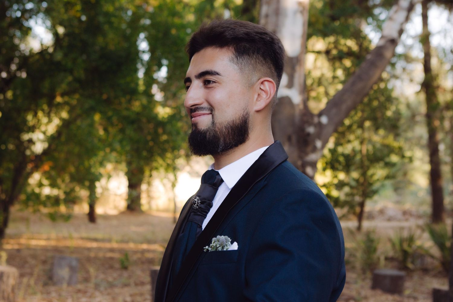 Stylish groom in navy suit with beard smiles confidently in a sunlit outdoor autumn setting.