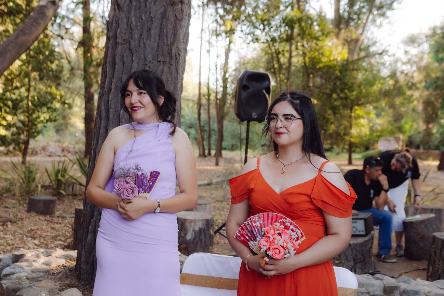 Two bridesmaids in lavender and red dresses holding bouquets at an outdoor wedding ceremony in a wooded setting.
