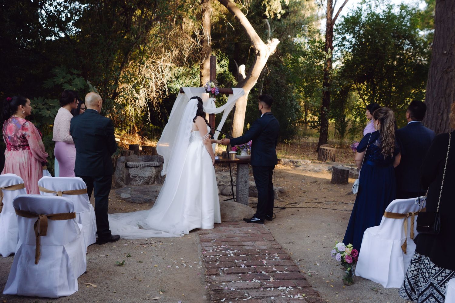 Bride and groom exchange vows at an elegant outdoor wedding ceremony surrounded by trees and white chairs.