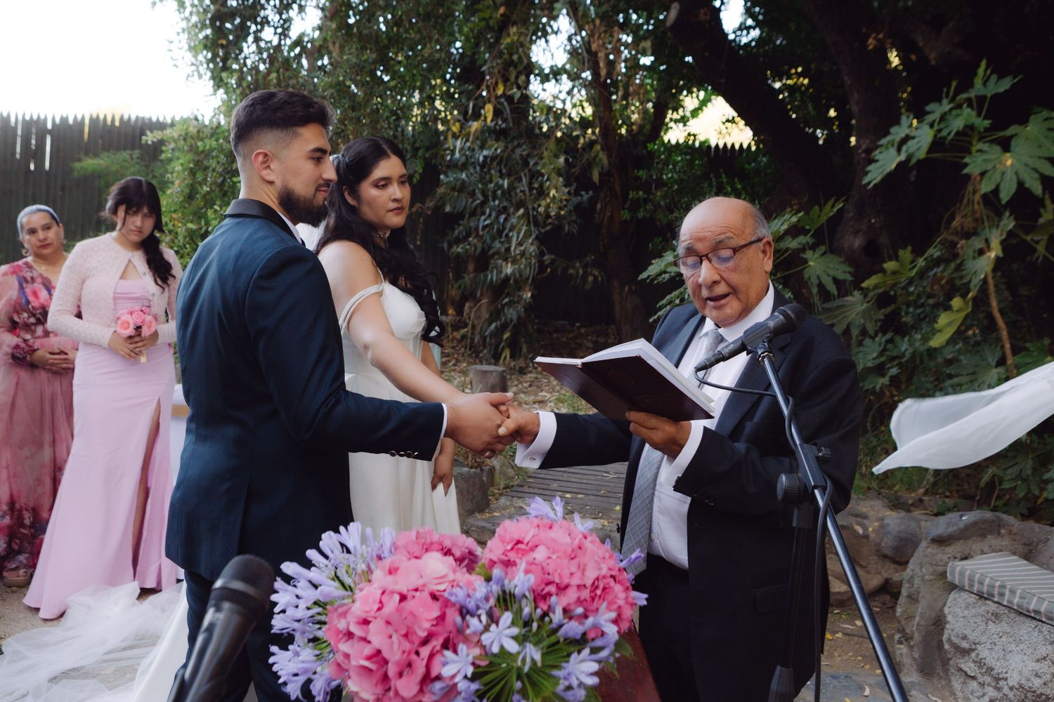 Officiant reads from a book as bride and groom hold hands during an outdoor garden wedding ceremony.