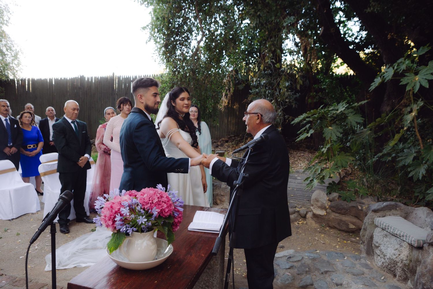 Groom shakes hands with elderly officiant at outdoor wedding ceremony beside a table with pink flowers.