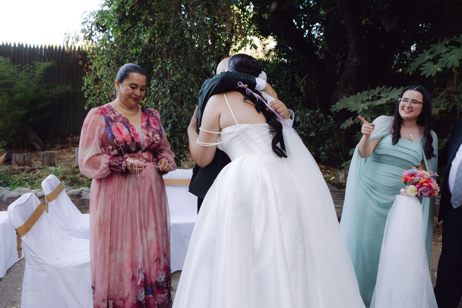 Bride in white gown embraces a guest in a floral dress at an outdoor wedding ceremony.