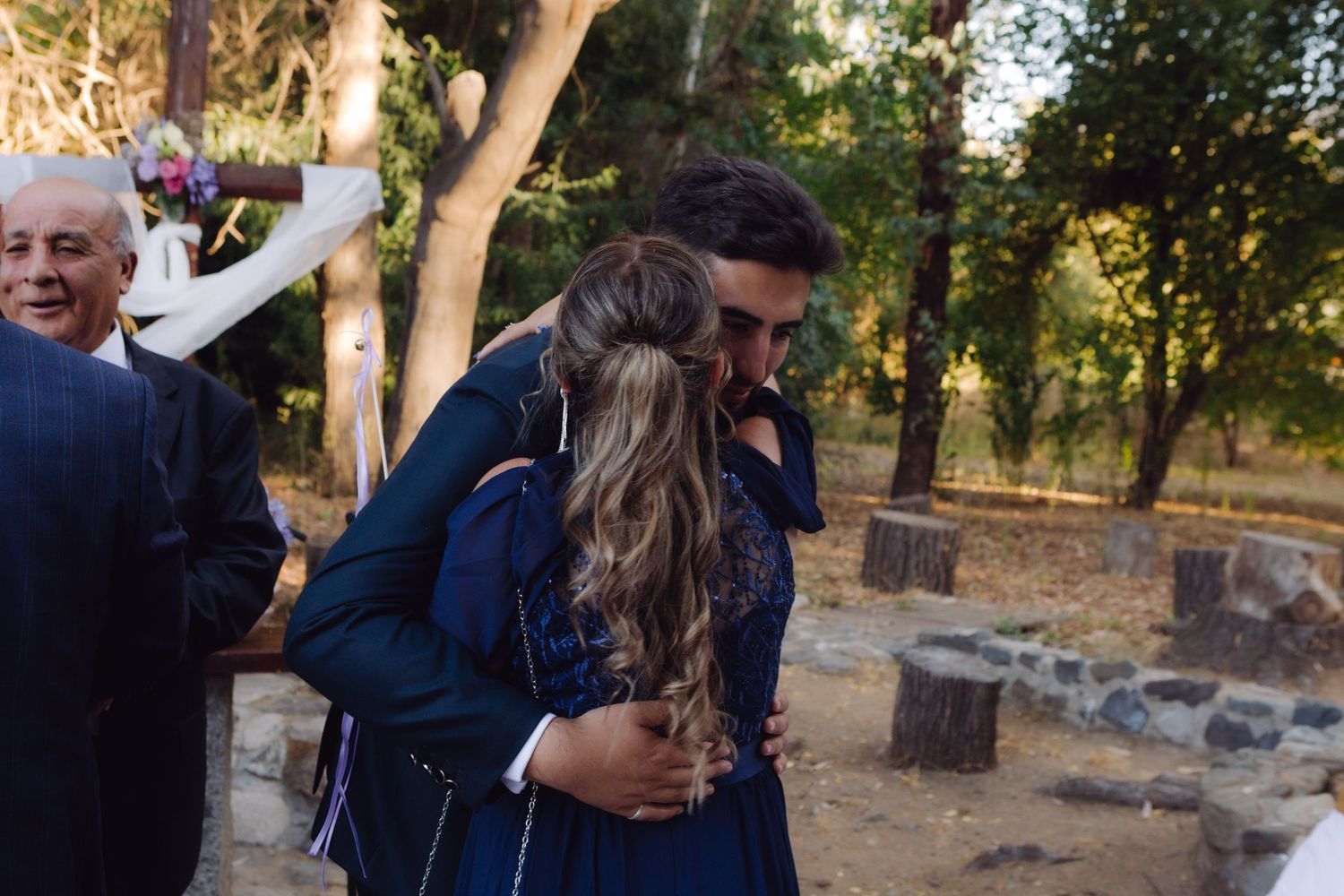 Groom in navy suit shares a warm embrace with a guest after an outdoor wedding ceremony.