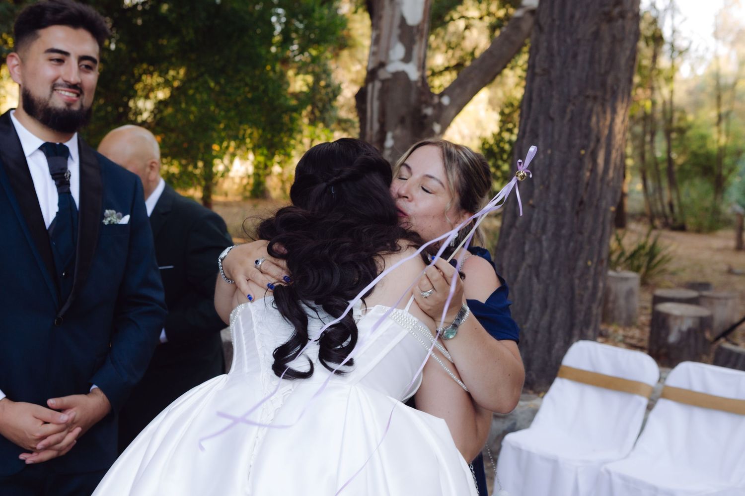 Bride in white wedding gown receives a heartfelt hug from a guest at an outdoor ceremony.