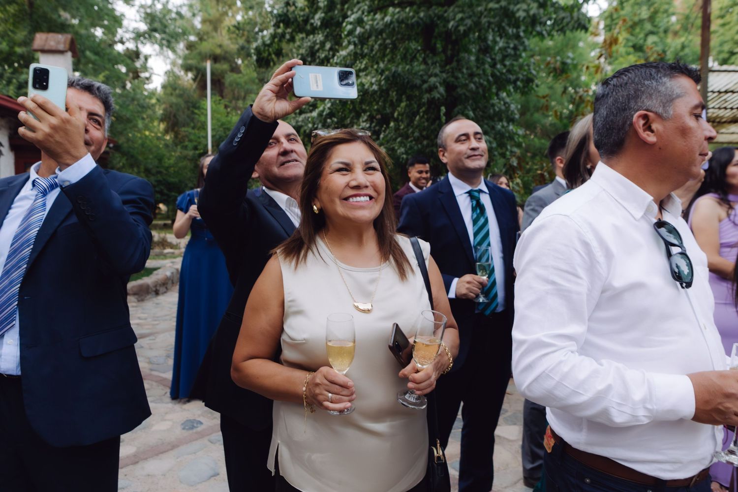 Wedding guests smile and take selfies outdoors while holding champagne glasses at a garden reception.