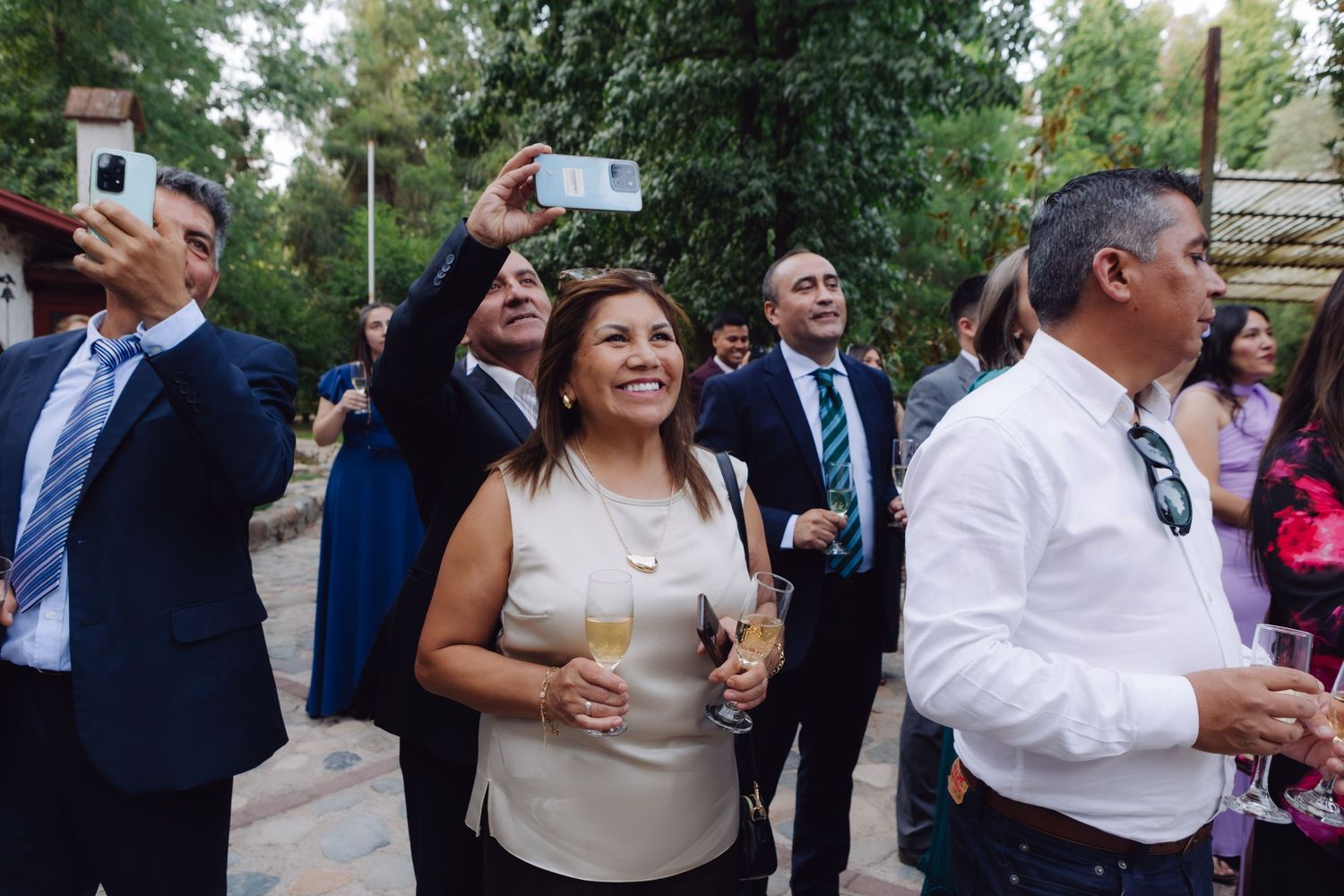 Cheerful wedding guests take photos and celebrate with champagne at an elegant outdoor garden reception.
