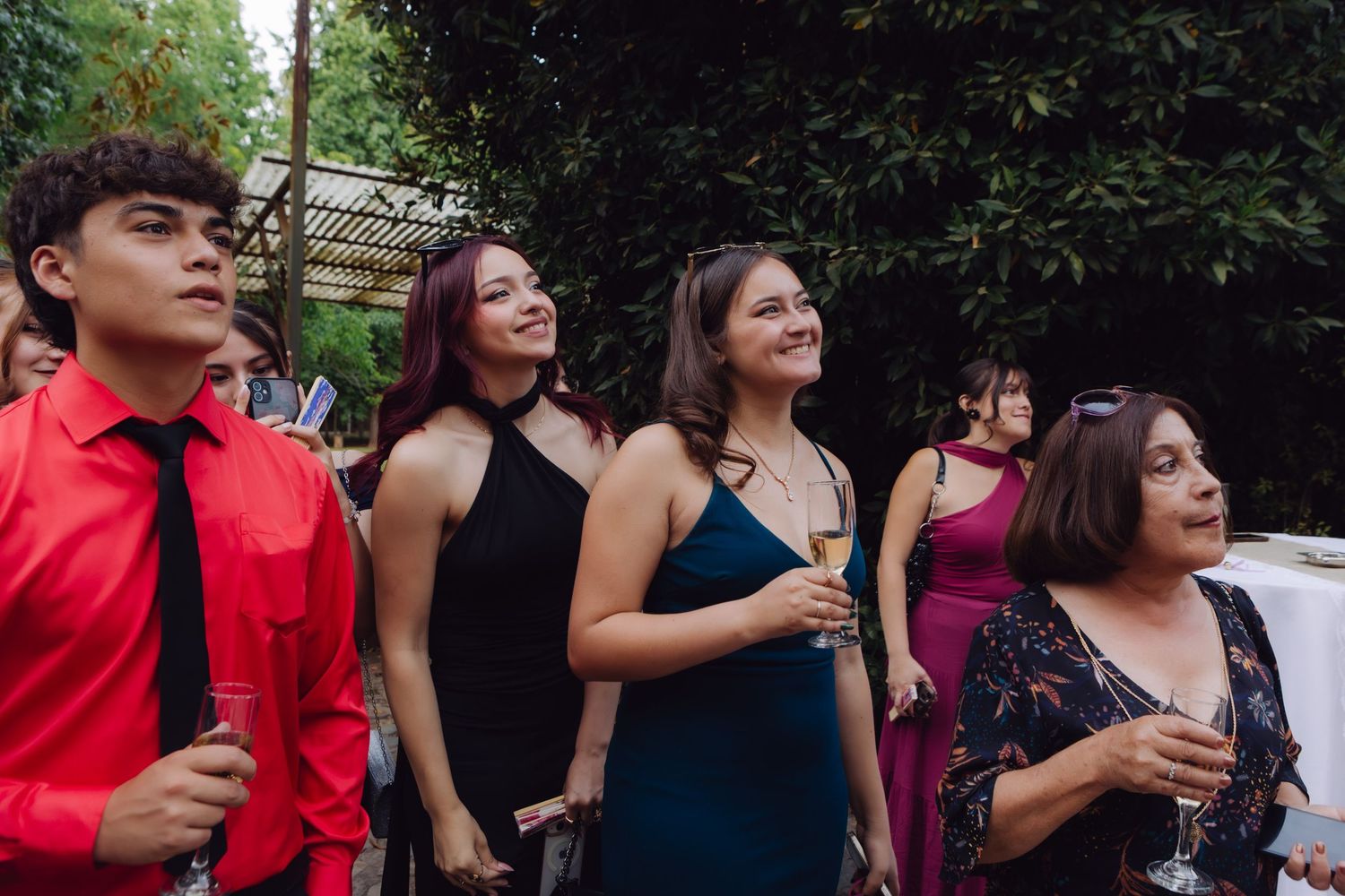 Elegantly dressed wedding guests smile and hold champagne glasses outdoors at a garden reception.