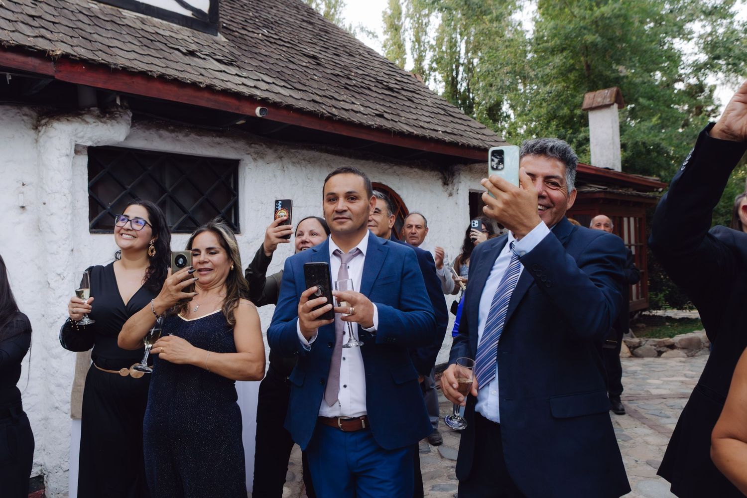 Smiling wedding guests in formal attire raise glasses and phones during an outdoor toast celebration.
