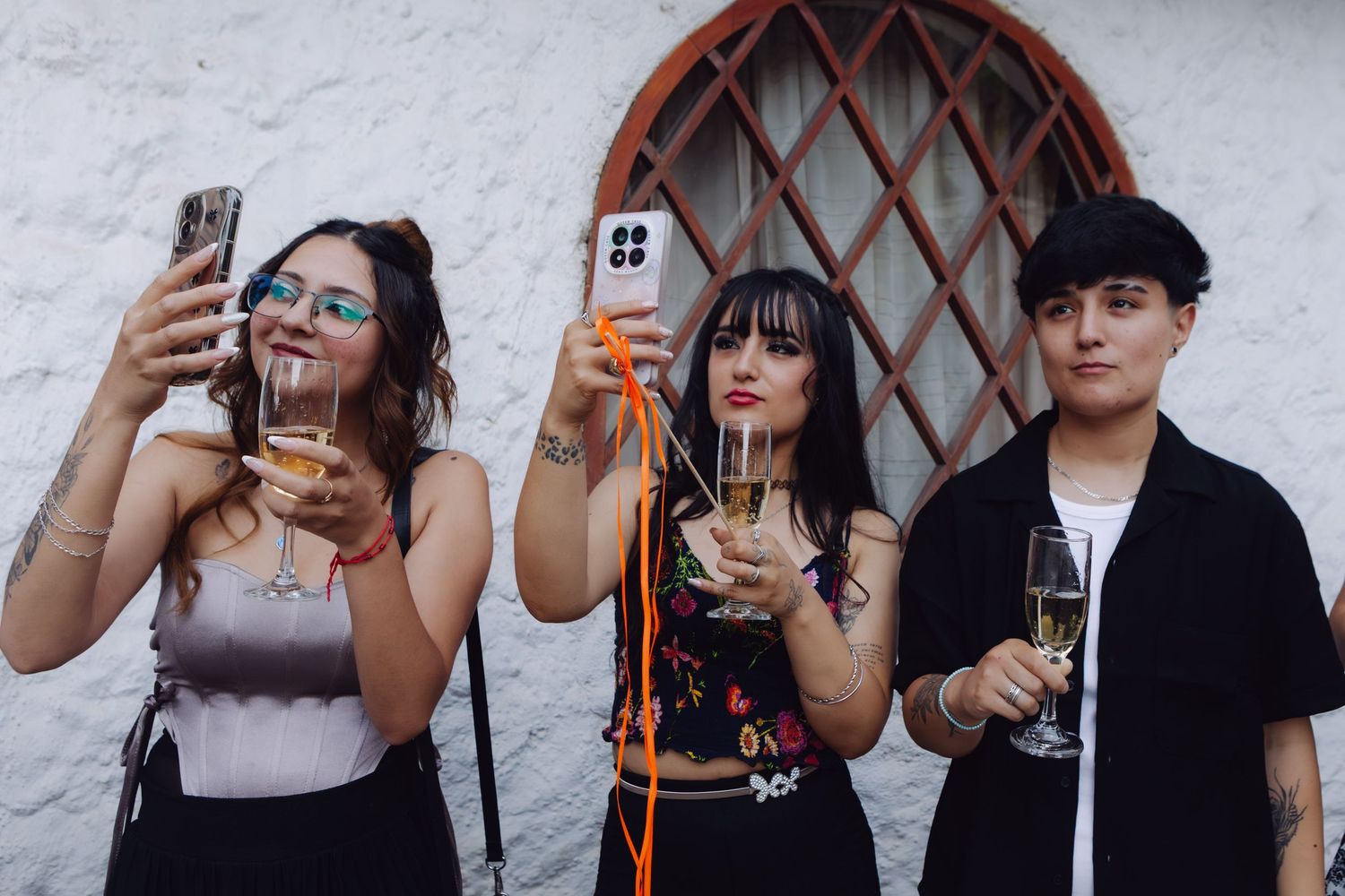 Three young guests pose with champagne glasses and phones at an outdoor wedding celebration.