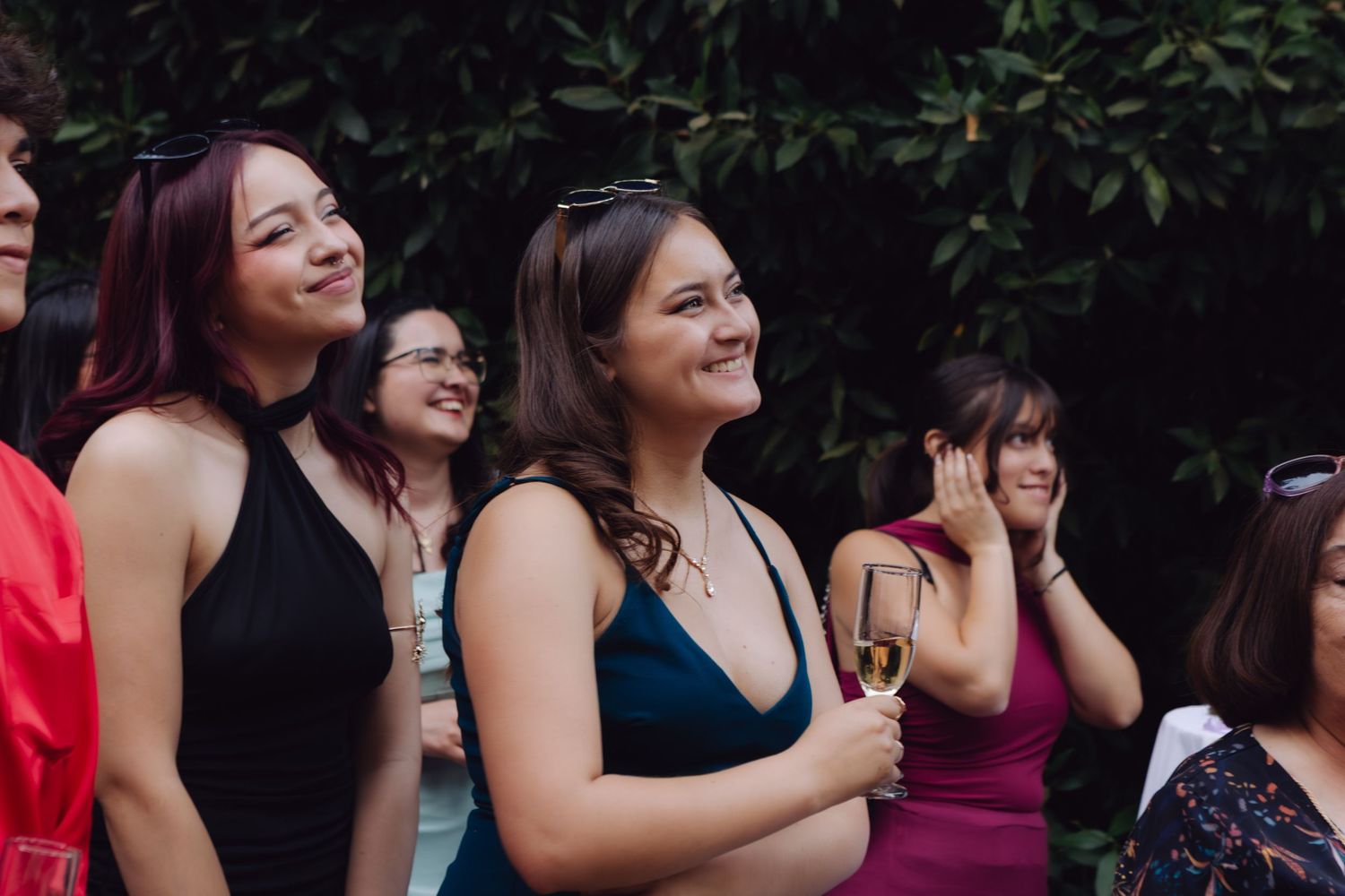 Two smiling women in formal dresses enjoy champagne at an outdoor wedding garden reception.