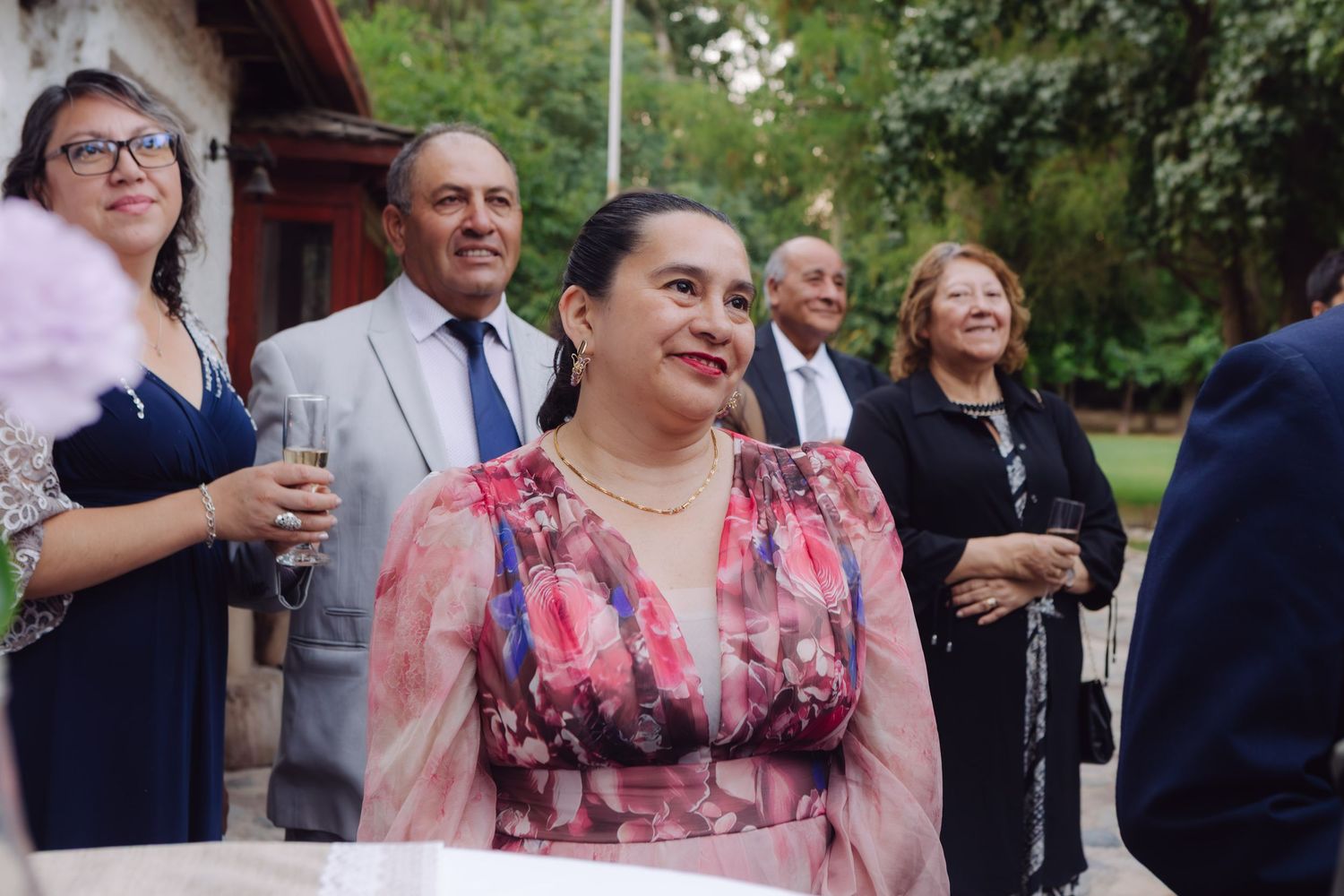 Woman in floral dress smiles warmly surrounded by formally dressed guests at outdoor wedding.