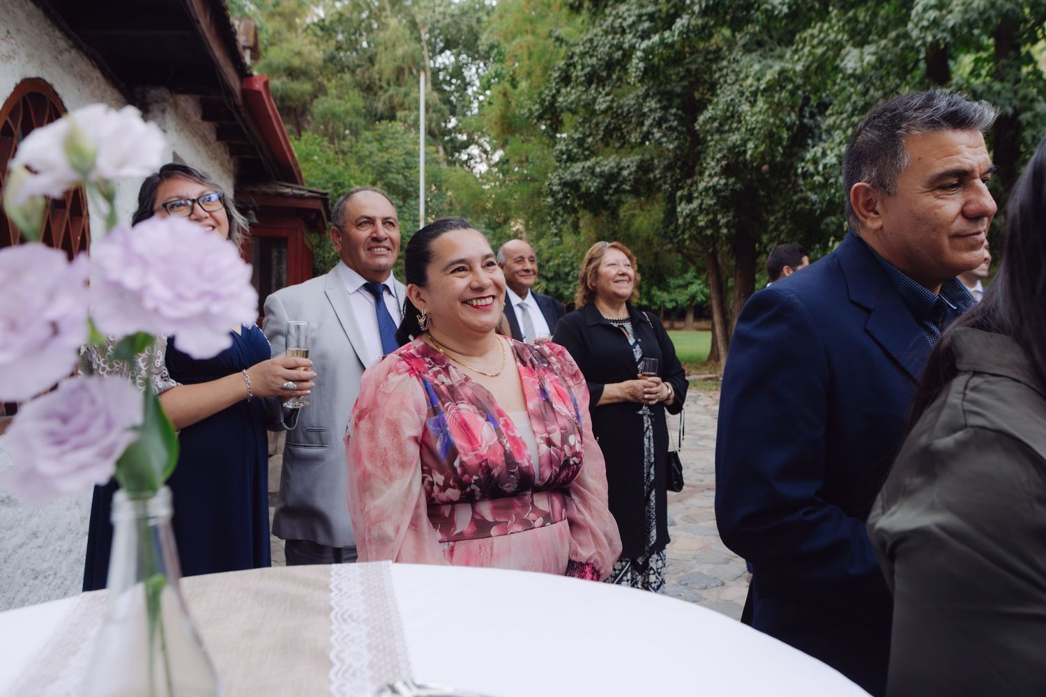 Elegantly dressed wedding guests smile and mingle at an outdoor reception with floral centerpieces.