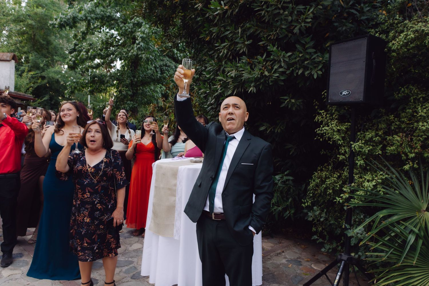 Man in tuxedo raises champagne glass in toast surrounded by festive wedding reception guests.