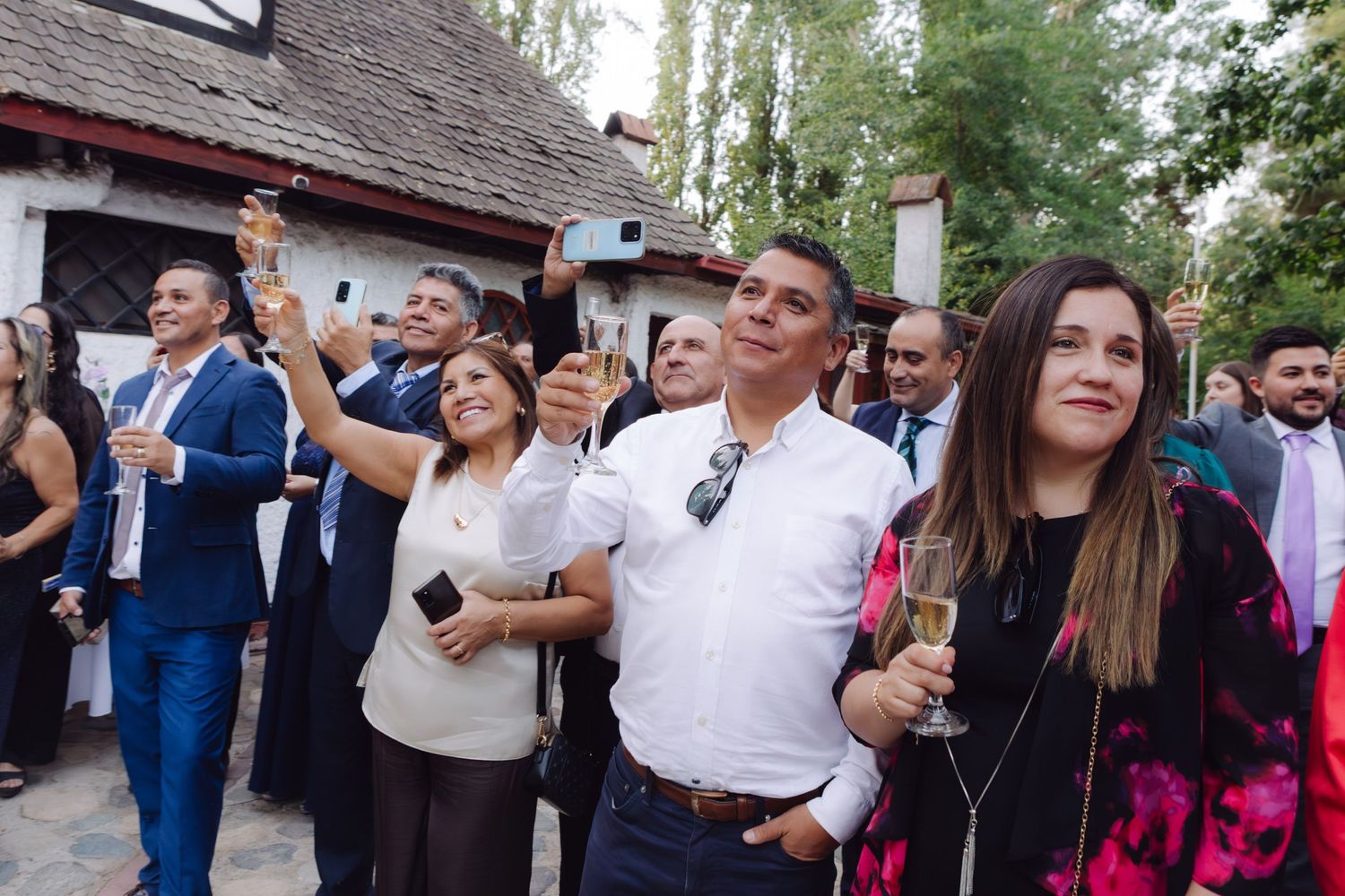 Smiling wedding guests raise champagne glasses during outdoor toast at elegant garden reception.