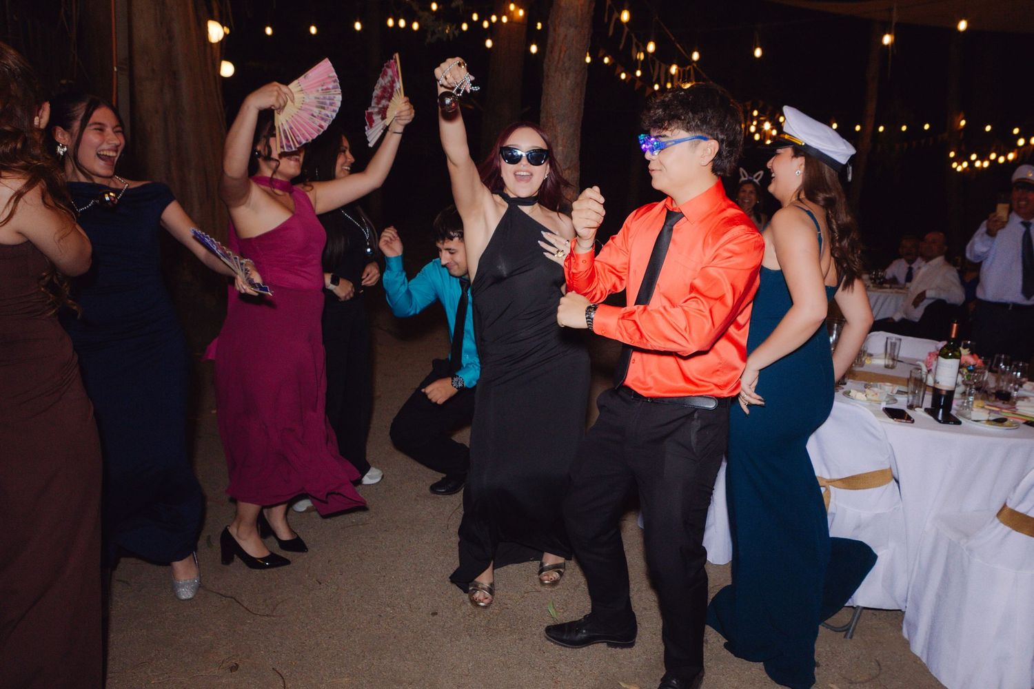 Energetic wedding guests dancing at outdoor reception under string lights at night.