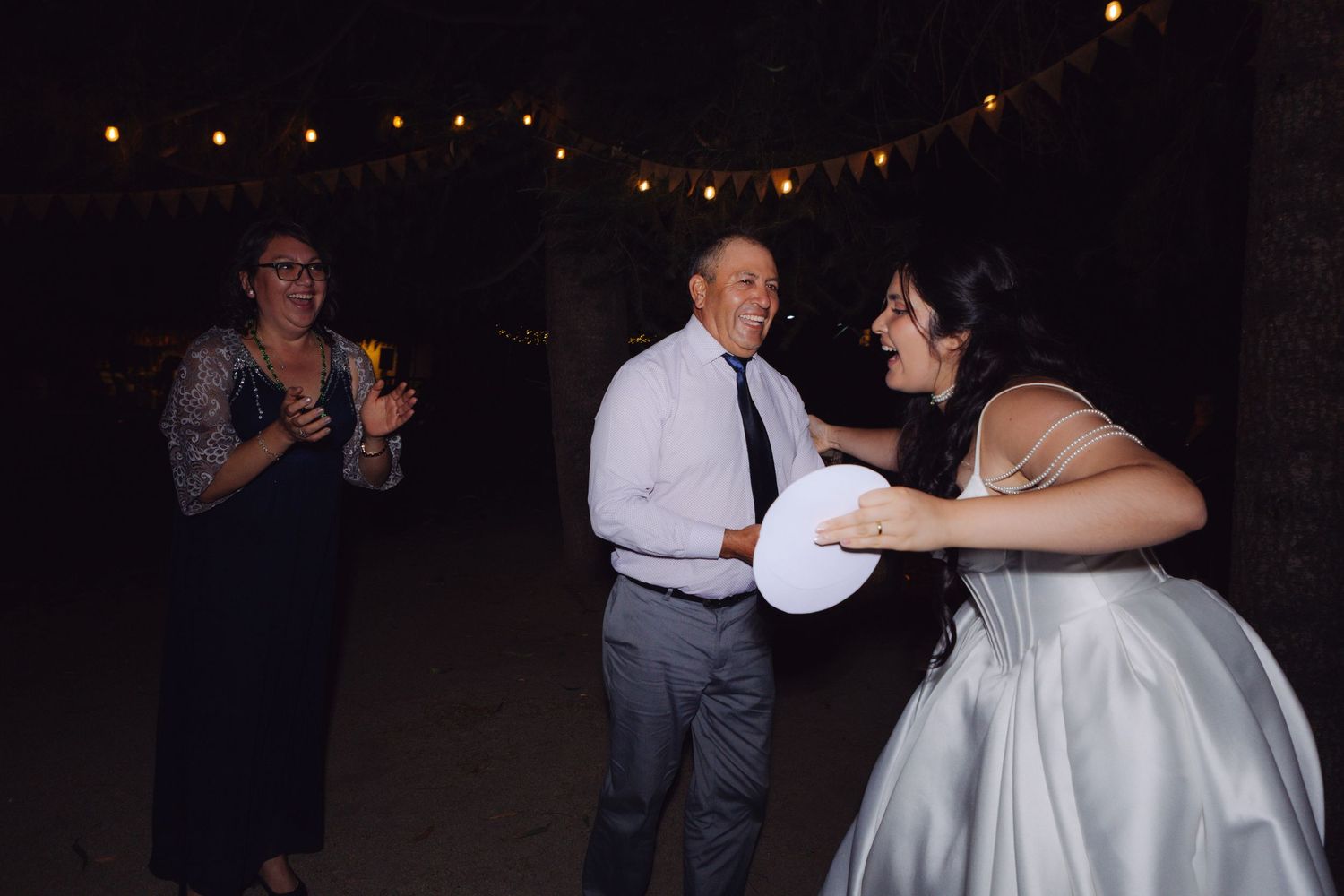 Bride and groom dancing joyfully with a guest at an outdoor nighttime wedding reception.