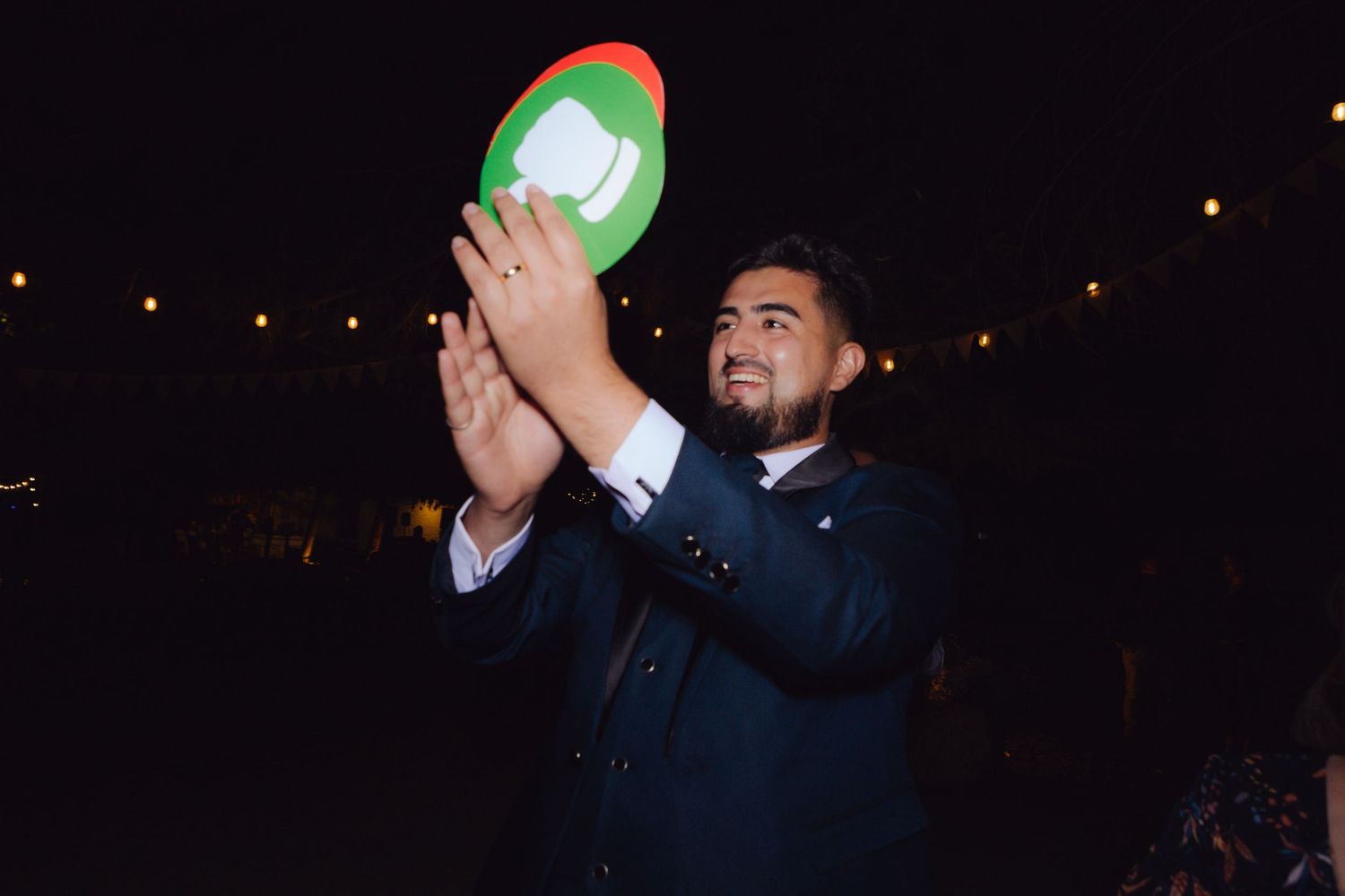 Man in suit holding a colorful sign at a festive nighttime wedding celebration event.