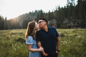 Couple sharing a kiss in a sunlit meadow with wildflowers and pine trees in the background.