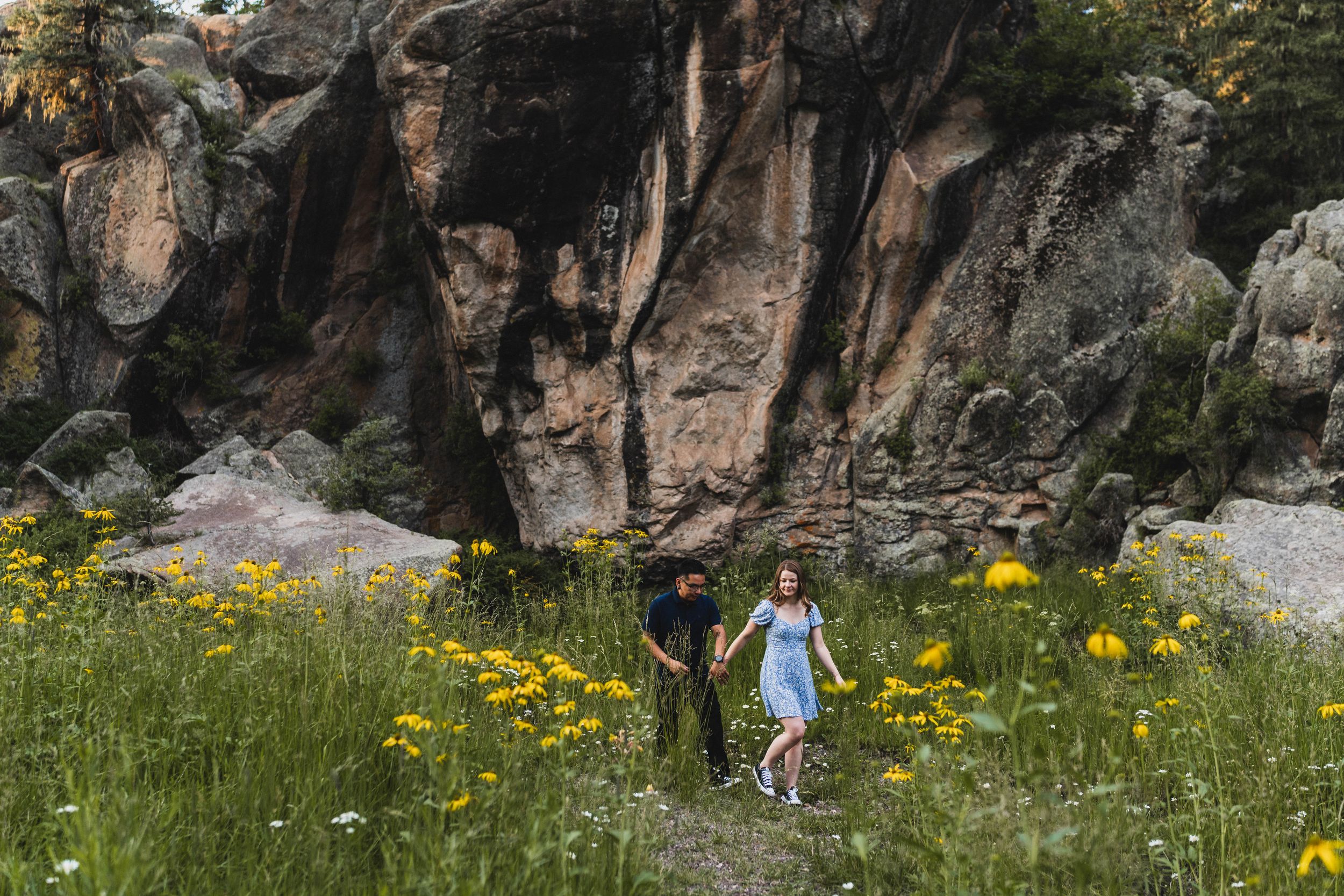 Couple holding hands walking through wildflower meadow with dramatic rocky cliffs in background.