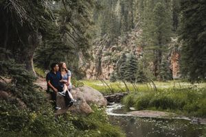 Couple sitting on rocks beside a serene mountain stream surrounded by lush pine trees and wildflowers in a forest setting.