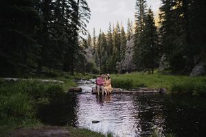 Couple sitting on logs in a serene mountain stream surrounded by tall pine trees at dusk.
