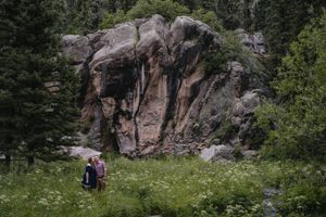 Two hikers stand before a massive red rock formation surrounded by lush green trees and wildflowers in a mountain forest.